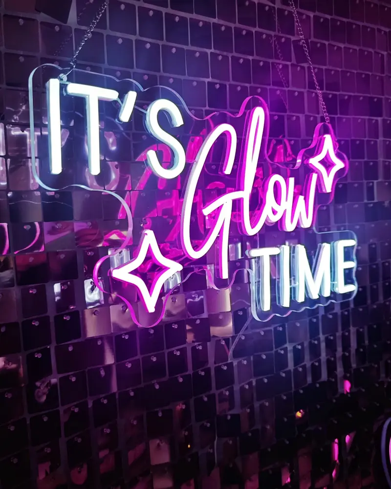 Cartel luminoso de neón «It’s Glow Time» del Glitter Bar sobre pared de lentejuelas